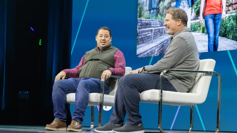 On the left, Rob Barlow sits on a chair. He wears a maroon shirt and green vest. On the right, Sam Johnson also sits on a chair. He wears dark pants and a green sweater. He is laughing.