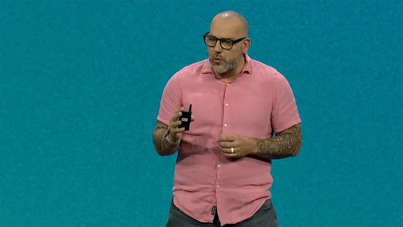 Mat Pullen has a shaved head and dark glasses. He stands in front of a teal JNUC 2025 background and gestures with his hands. He wears a short-sleeved pink dress shirt, and has black tattoos on each of his forearms.