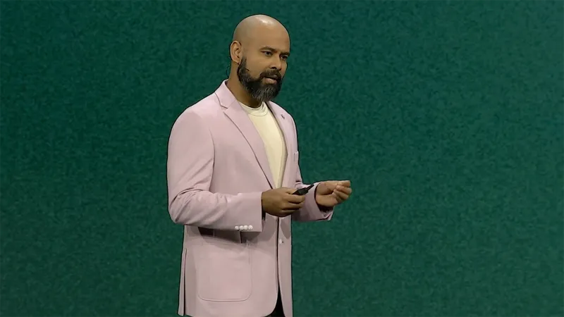 Suraj Mohandas presents from the stage at JNUC 2025. He has a closely trimmed beard and wears a lavender suit jacket.