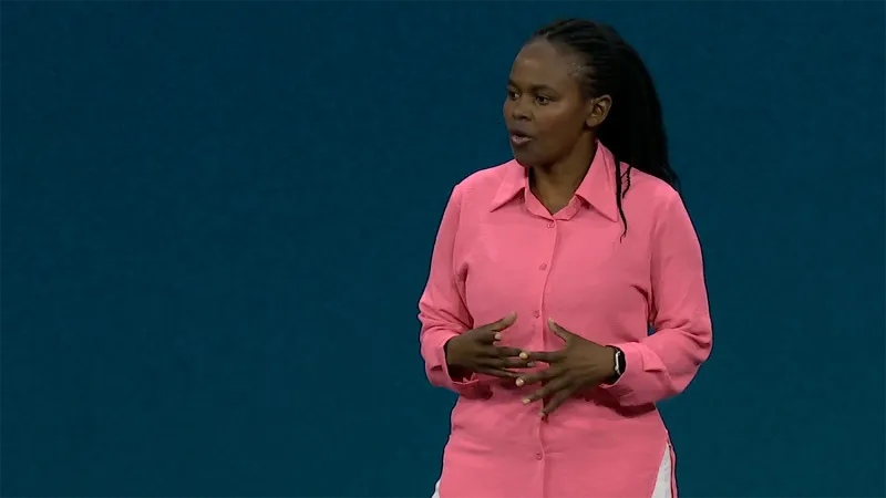 Tsitsi-Murohoke stands in front of a dark blue JNUC 2025 backgrounds. She makes a small gesture with her hands and looks slightly to the left. She wears her long hair in small, tight braids pushed back from her face. She wears a bright pink dress shirt.