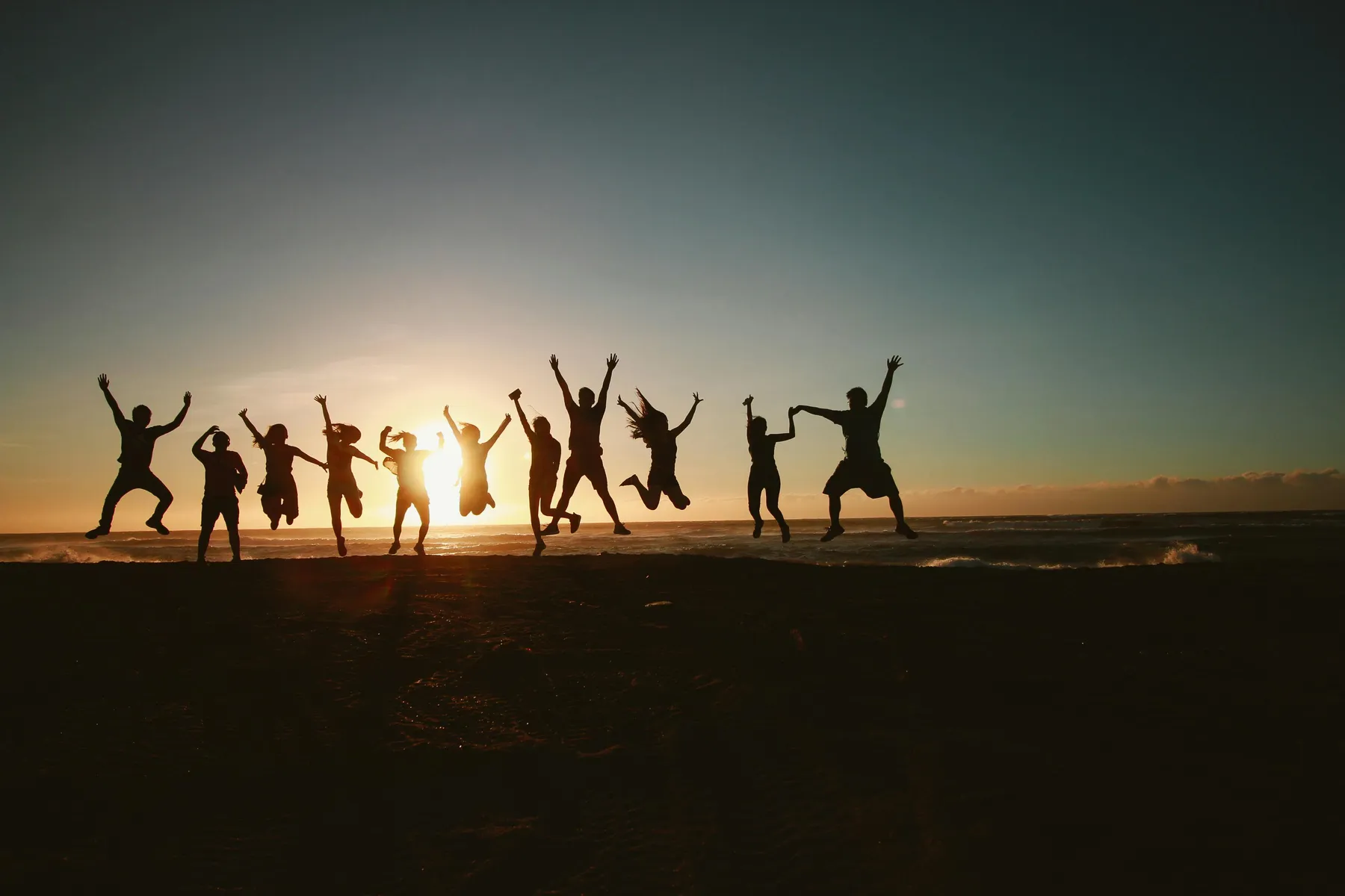 A group of people on a beach, backlit by the sun rising over the water, jump into the air