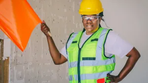 A person in an orange hardhat, safety vest, and safety goggles holds up an orange card to symbolize safety compliance.