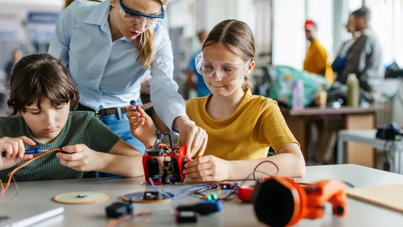 A teacher assists two students with building a robotics robot in her active learning classroom. They wear safety classes.