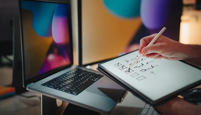 Hands holding up a checklist and pen in front of a Mac to illustrate automating macOS compliance implementation, maintenance, and enforcement with Jamf.