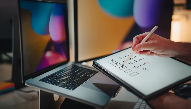 Hands holding up a checklist and pen in front of a Mac to illustrate automating macOS compliance implementation, maintenance, and enforcement with Jamf.