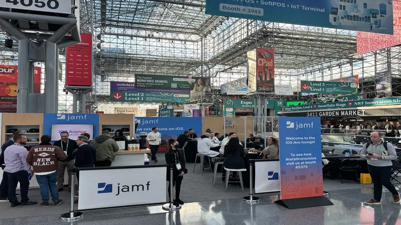 Jamf booth and iOS Ave lounge at NRF 2026 trade show, with attendees gathered around demo counters and seating under a large convention hall ceiling with directional signage overhead.