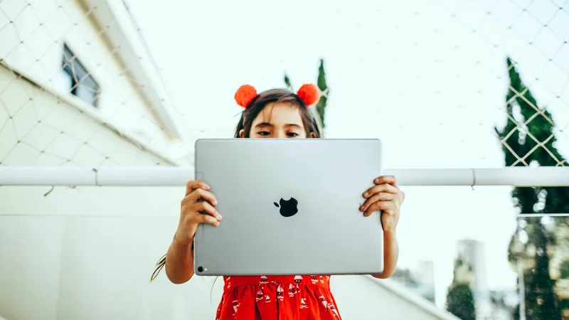 A young student in a bright red dress peeks over the edge of an iPad she holds upraised.