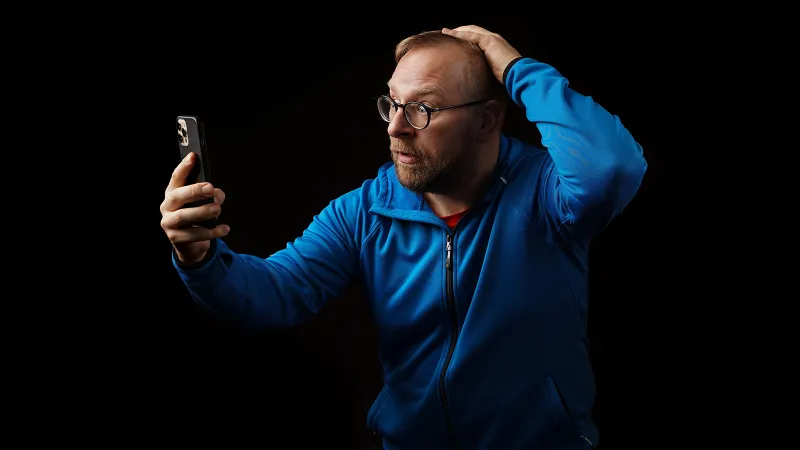 Illustration of mobile phishing: A pale-skinned man wearing a blue shirt looks into a mobile phone in shock and pulls at his hair.