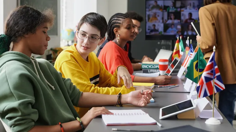Diverse group of students talking at a long desk with tablets, notebooks, and international flags in a classroom.