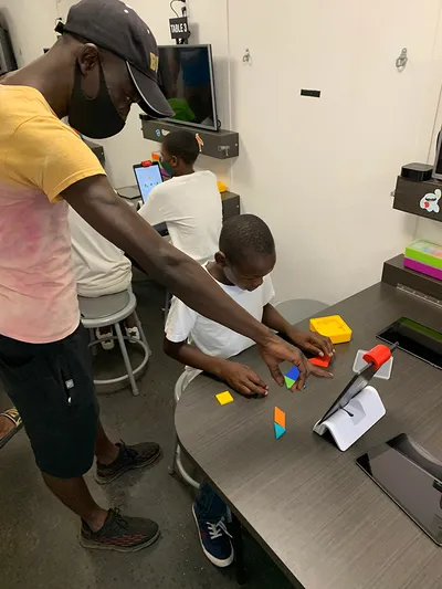 Three students and a teacher at a Zimbabwe MATTER Innovation Hub hard at work. The teacher points at an iPad propped up in front of a student in a white T-shirt, whose gaze is focused intently at colorful shaped he is putting together. Two other students