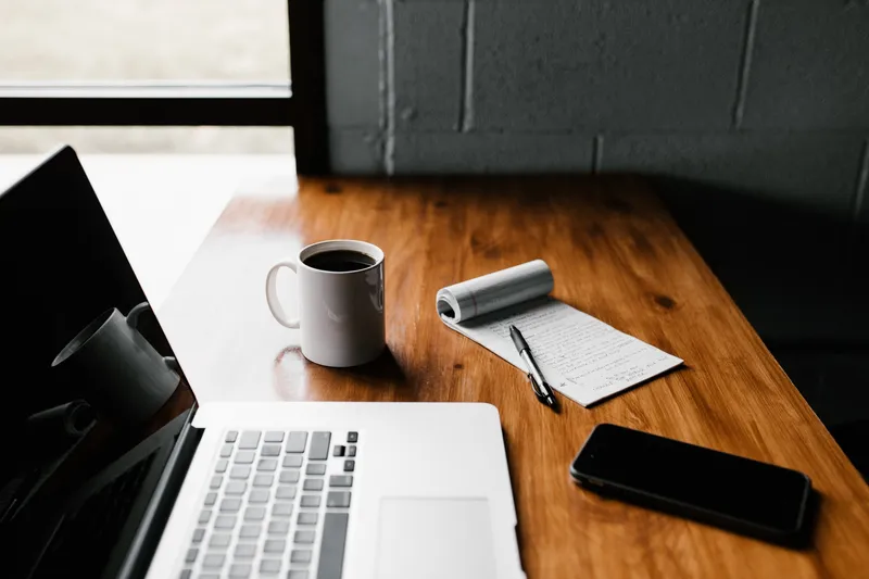 MacBook, pad of paper, cup of coffee and an iPhone sitting on a desk.