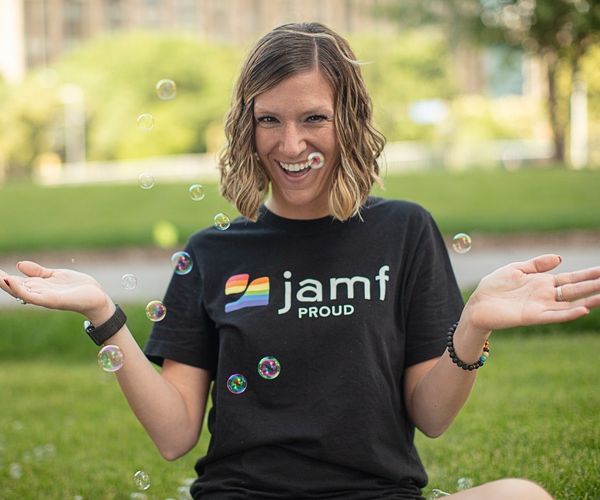 Woman in Jamf Proud t-shirt sits on grass in a park, smiling, arms spread behind bubbles floating in the air.