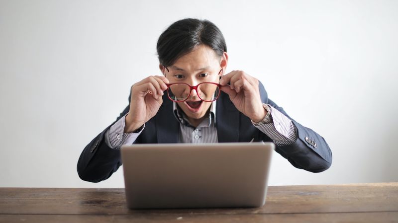 Man adjusts glasses on face while looking in surprise at computer screen.