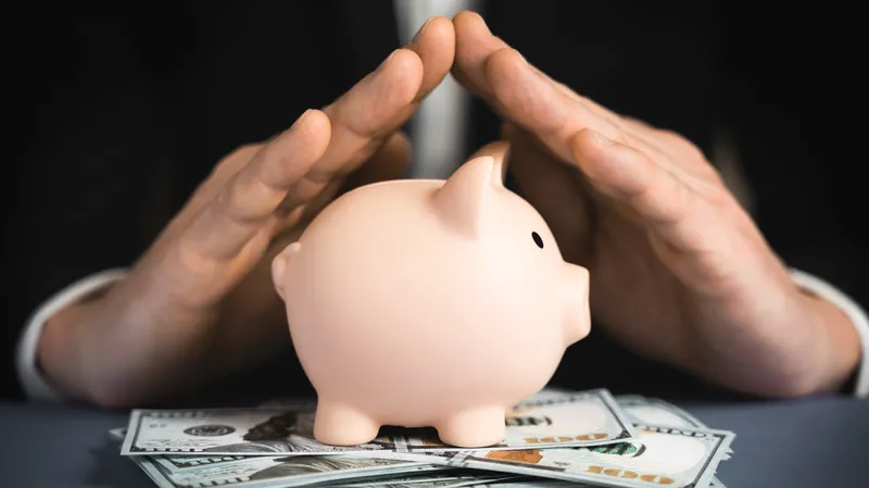Money and a piggy bank on a table with hands held together forming a small roof, symbolizing protection of funds