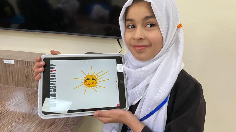 A young MATTER Innovation Hub student in Afghanistan holds up her iPad drawing of a bright yellow sun. She wears a black dress, a white headscarf and a proud smile.