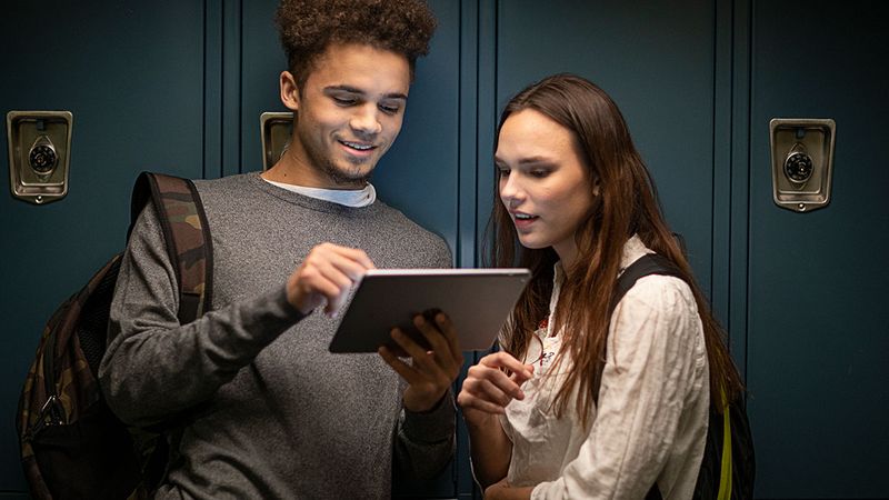 Two high school students stand in front of lockers and look at the same iPad together.