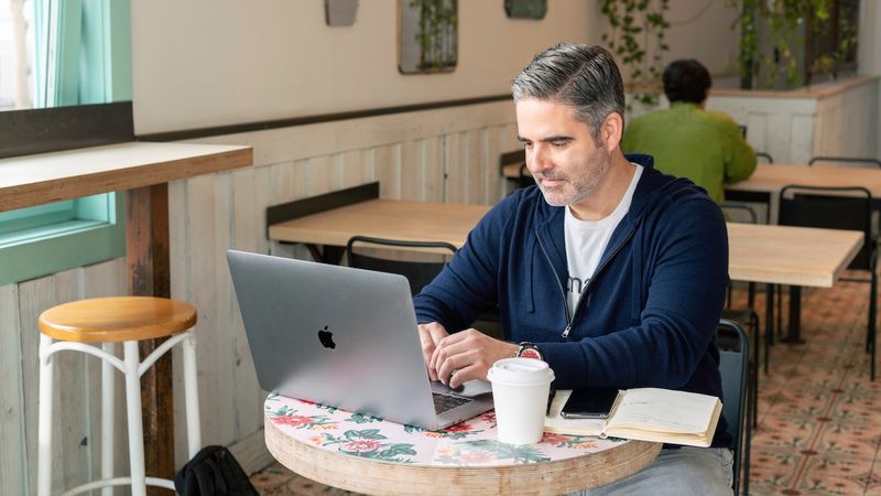 Man sits at a table in a coffee shop working on a MacBook managed by Jamf with Zero Trust Network Access.