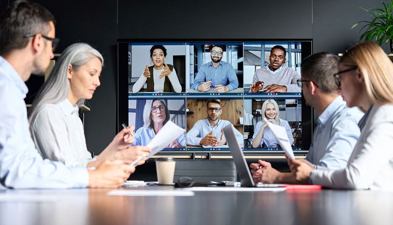 Group of employees at a conference table, collaborating online with remote users via webcam.