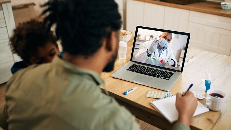 A father with a sick child on his shoulder uses Apple telehealth to speak with his doctor on a laptop.