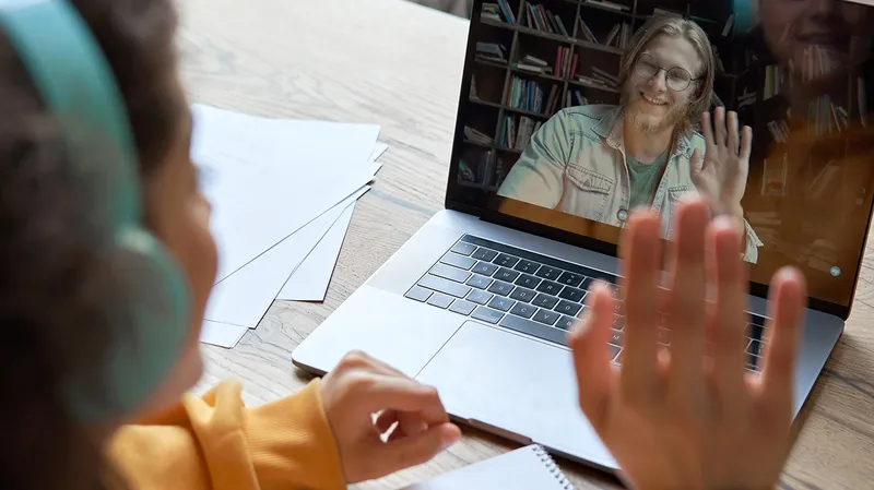 student wearing headphones, waving to someone on a MacBook screen