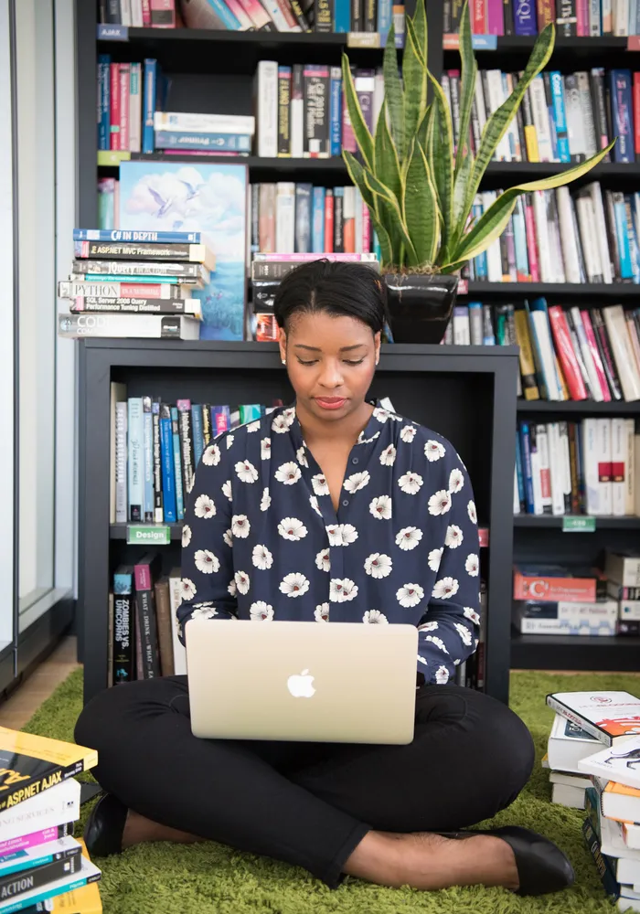 Woman sits cross-legged in a library, surrounded by books and using a MacBook.