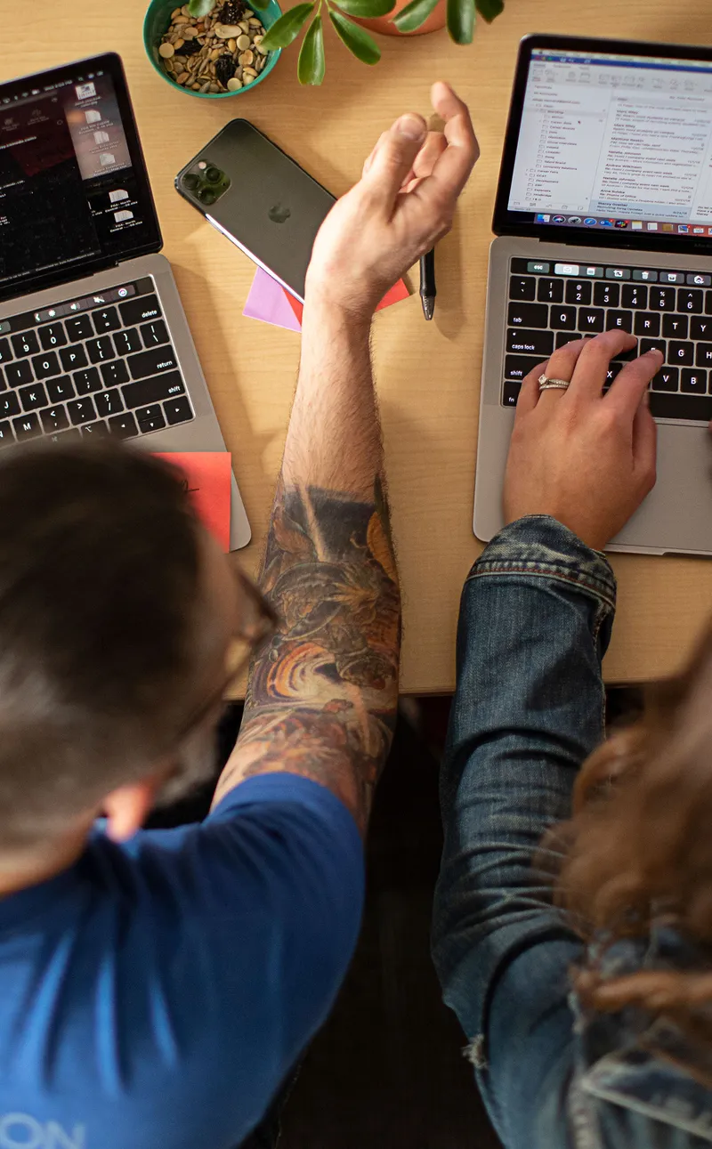 Two people sitting at a desk with MacBooks and an iPhone