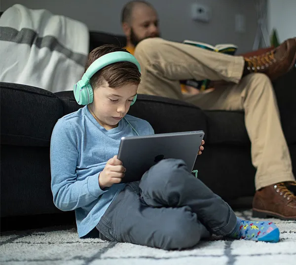 A child wearing headphones sits on a living room floor doing homework on a Jamf-managed iPad while his parent supervises.