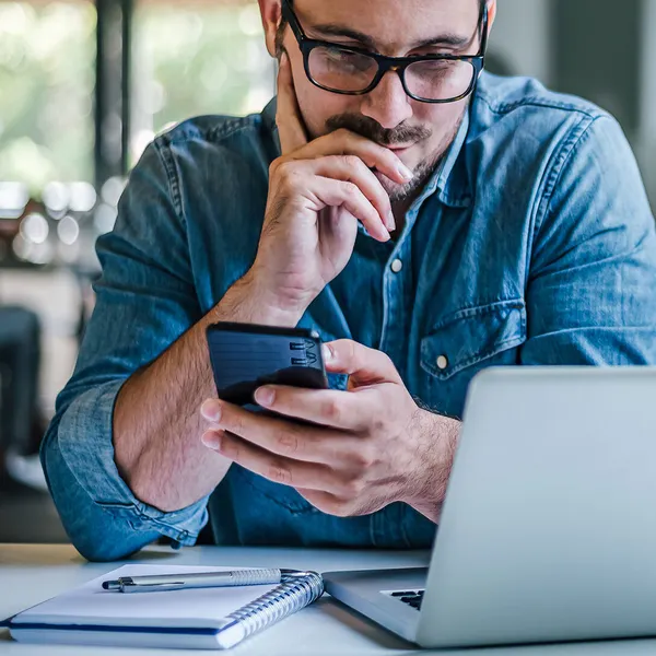 Male in a denim button up shirt with beard and glasses is using an Apple iPhone and Macbook at a desk.