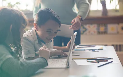 Young students smiling while using an iPad at a classroom desk, with a teacher nearby and colored pencils spread across the table.