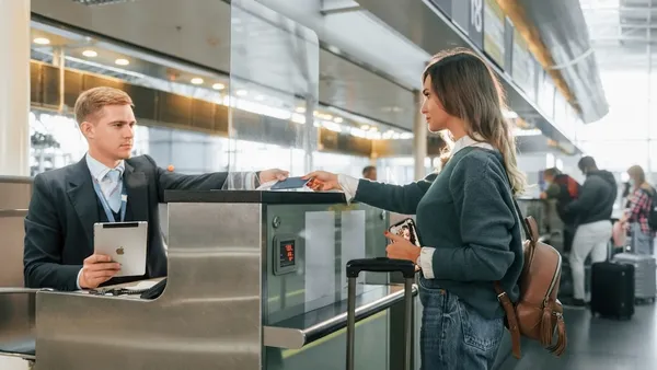 A female airline passenger speaks to another woman at the ticketing gate counter of a large airport.