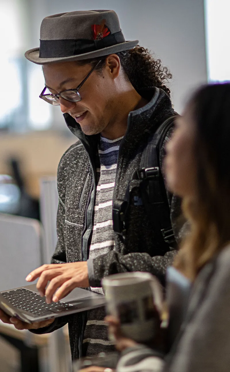 Man holding a MacBook with a woman foregrounded, holding a cup, but blurred from vision